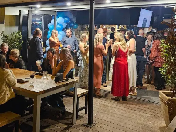 A lively gathering at an outdoor bar with people socializing, drinks on tables, and blue balloons in the background.